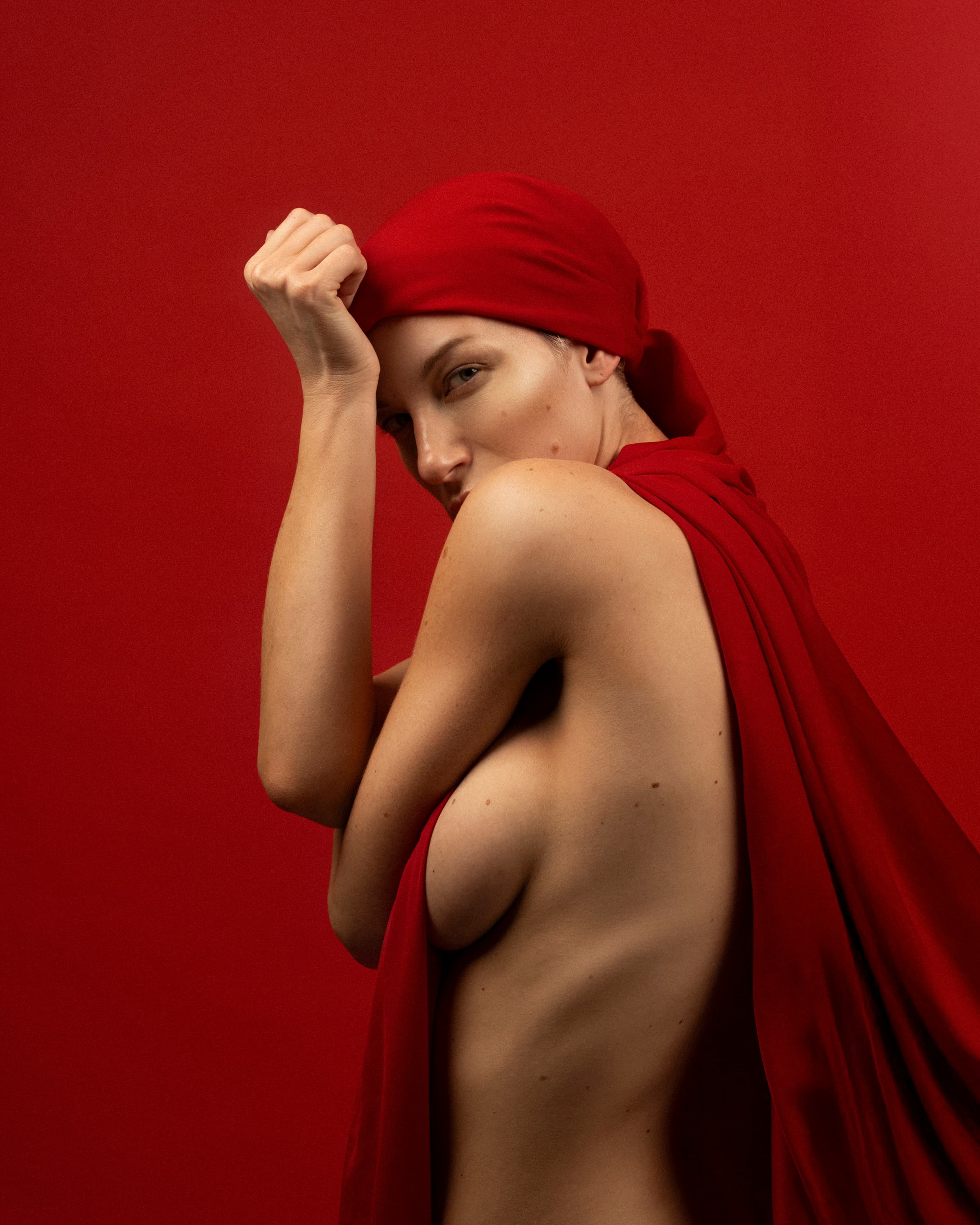 A woman wrapped in red fabric against a red backdrop, looking over her shoulder