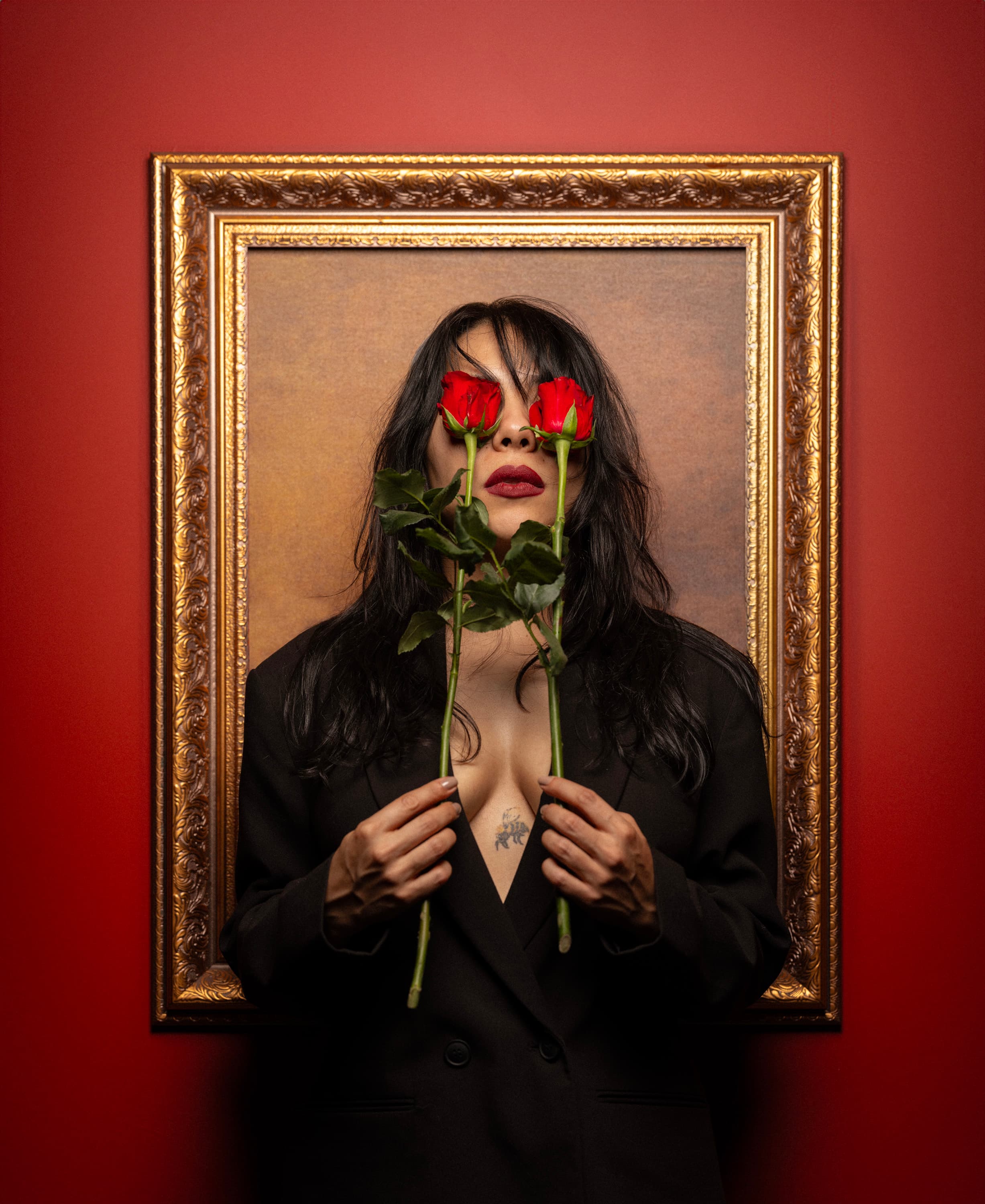 A woman inside a gilded frame on a red wall, holding two red roses over her eyes