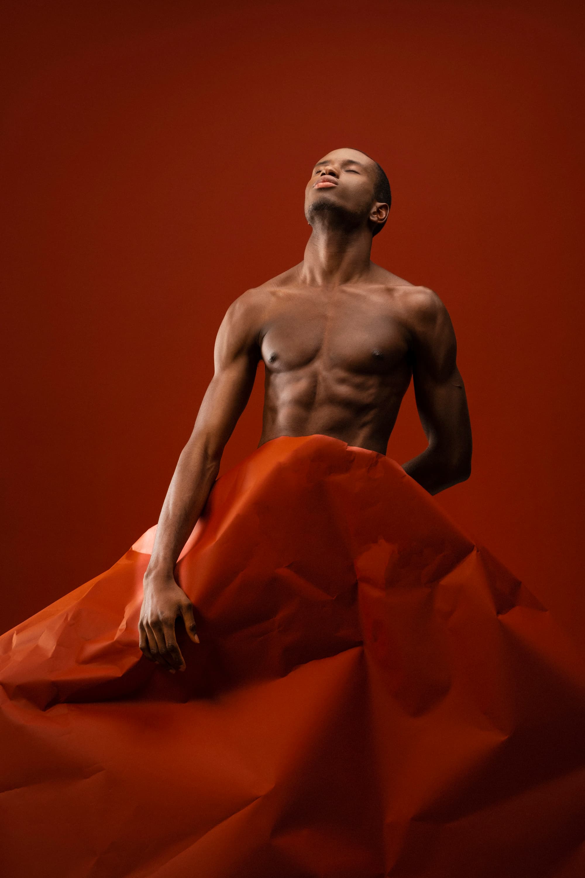 A shirtless man draped in rust-orange paper against a matching backdrop, head tilted back