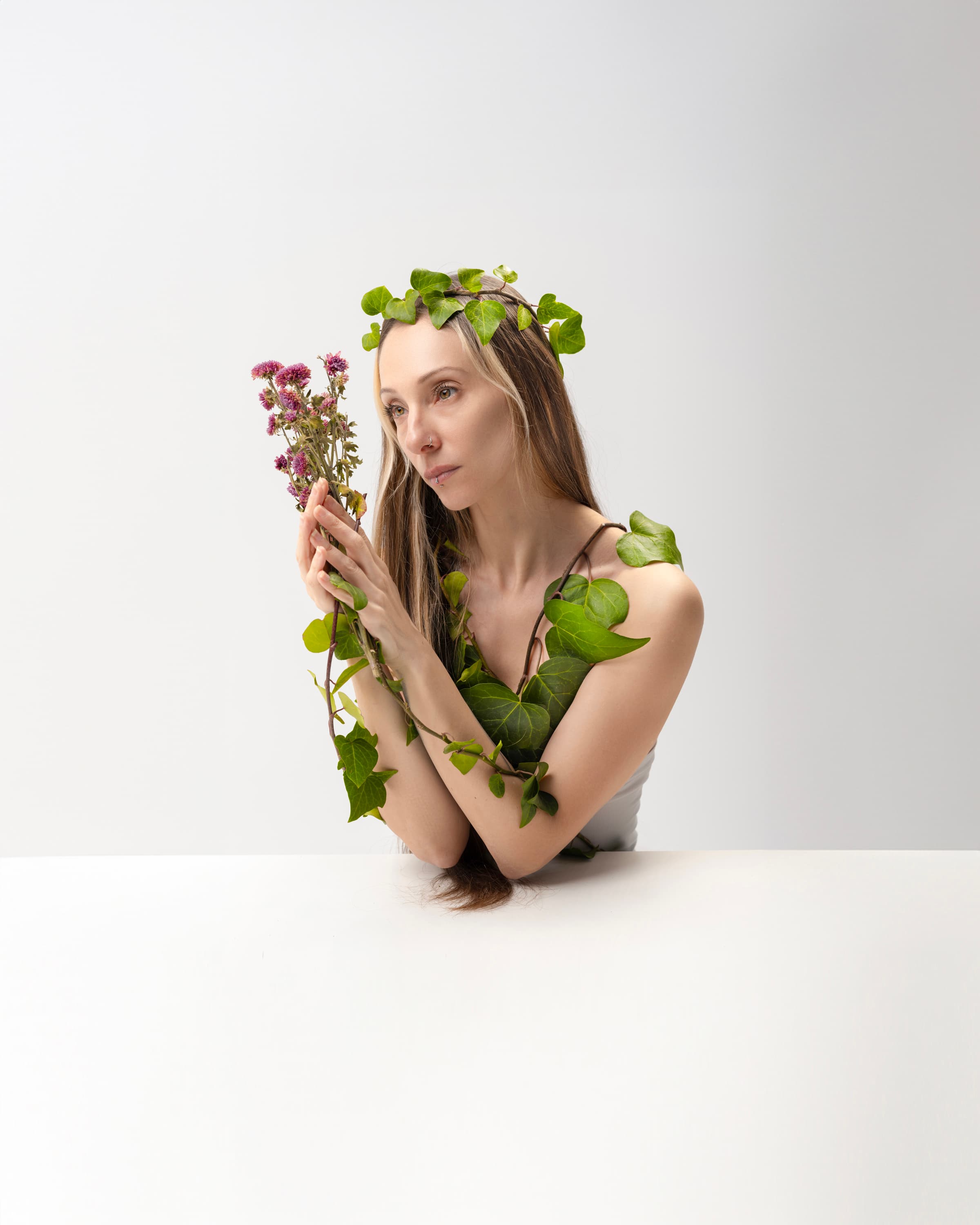 A woman with a leaf crown holds pink flowers at a white table, ivy wrapped around her arms