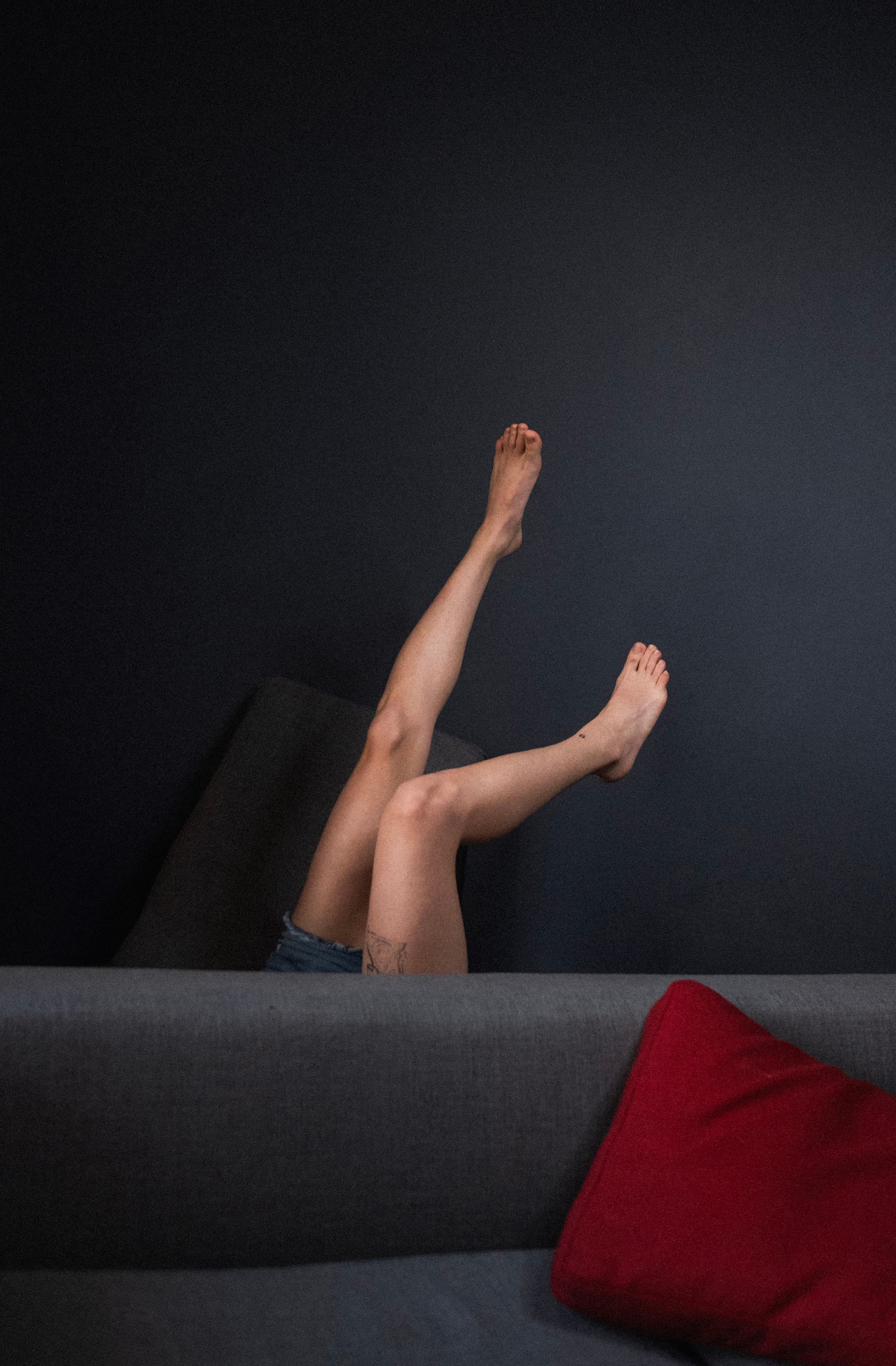 A pair of legs raised against a dark wall above a grey sofa with a red cushion