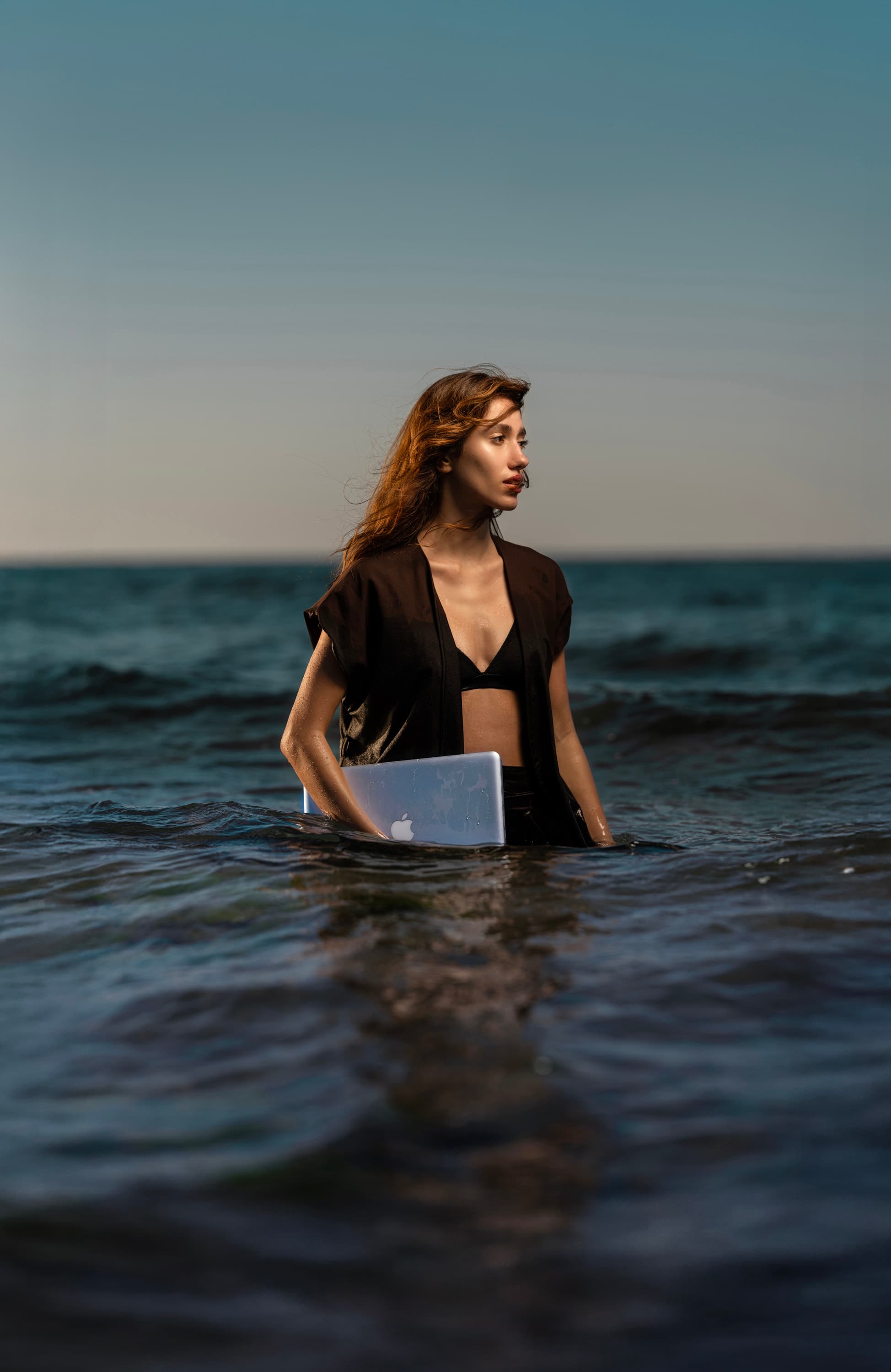 A woman stands waist-deep in the sea holding a laptop