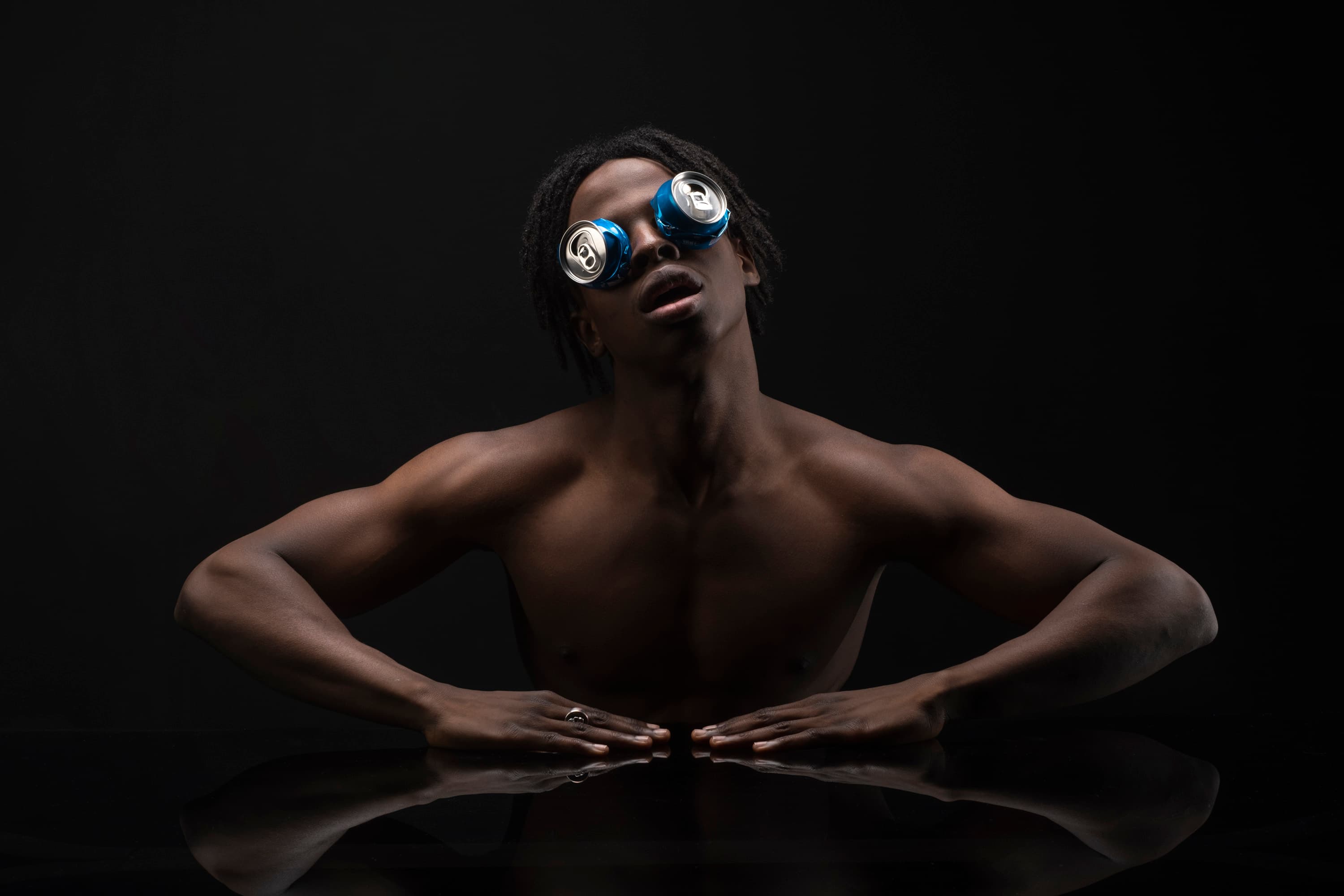 A shirtless man with cans over his eyes rests his hands on a reflective black table