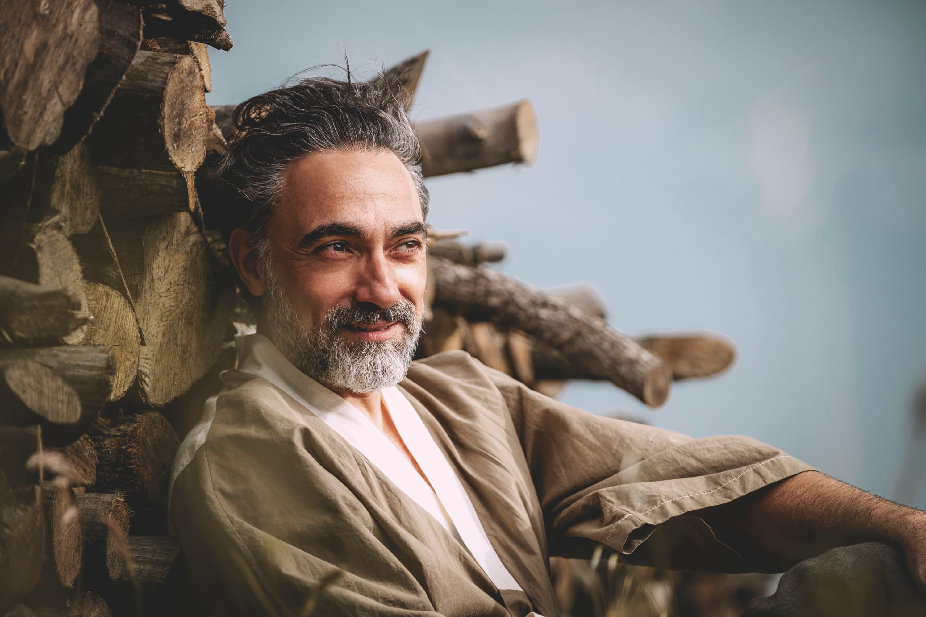 A bearded man in a linen jacket smiling in front of stacked logs