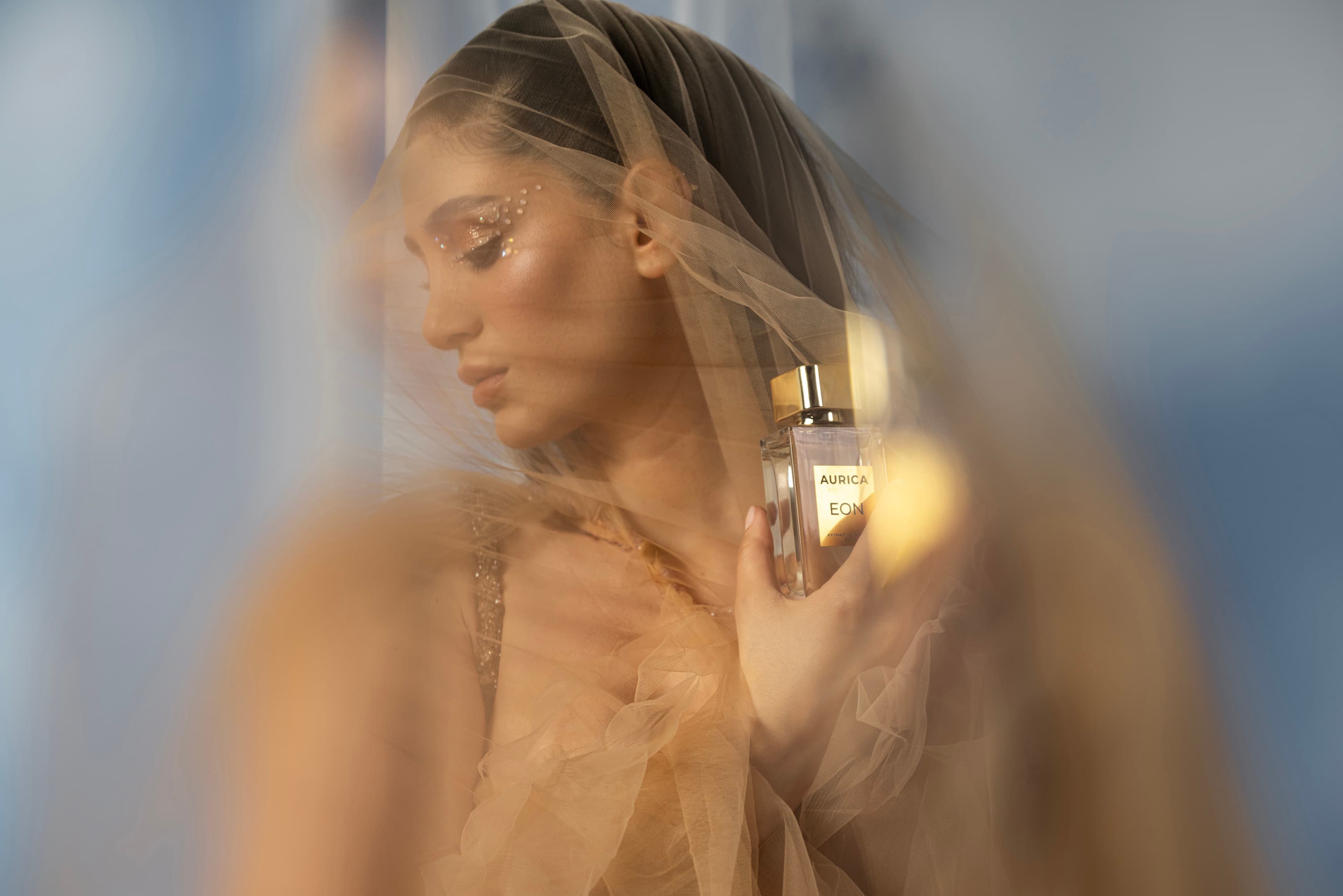 A woman in a soft tulle veil holds a perfume bottle in warm diffused light