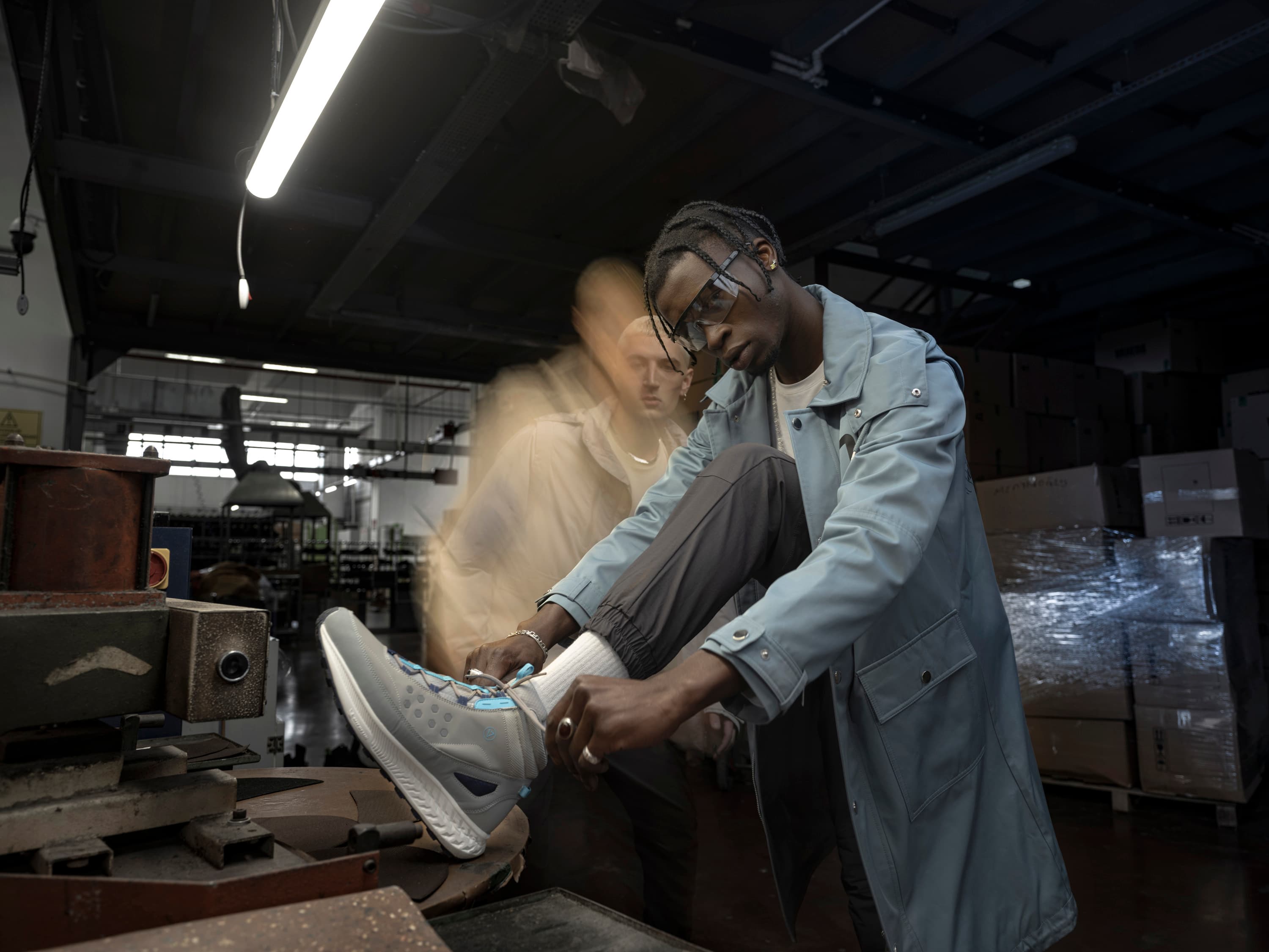A man in a light jacket tying his sneaker in an industrial workshop