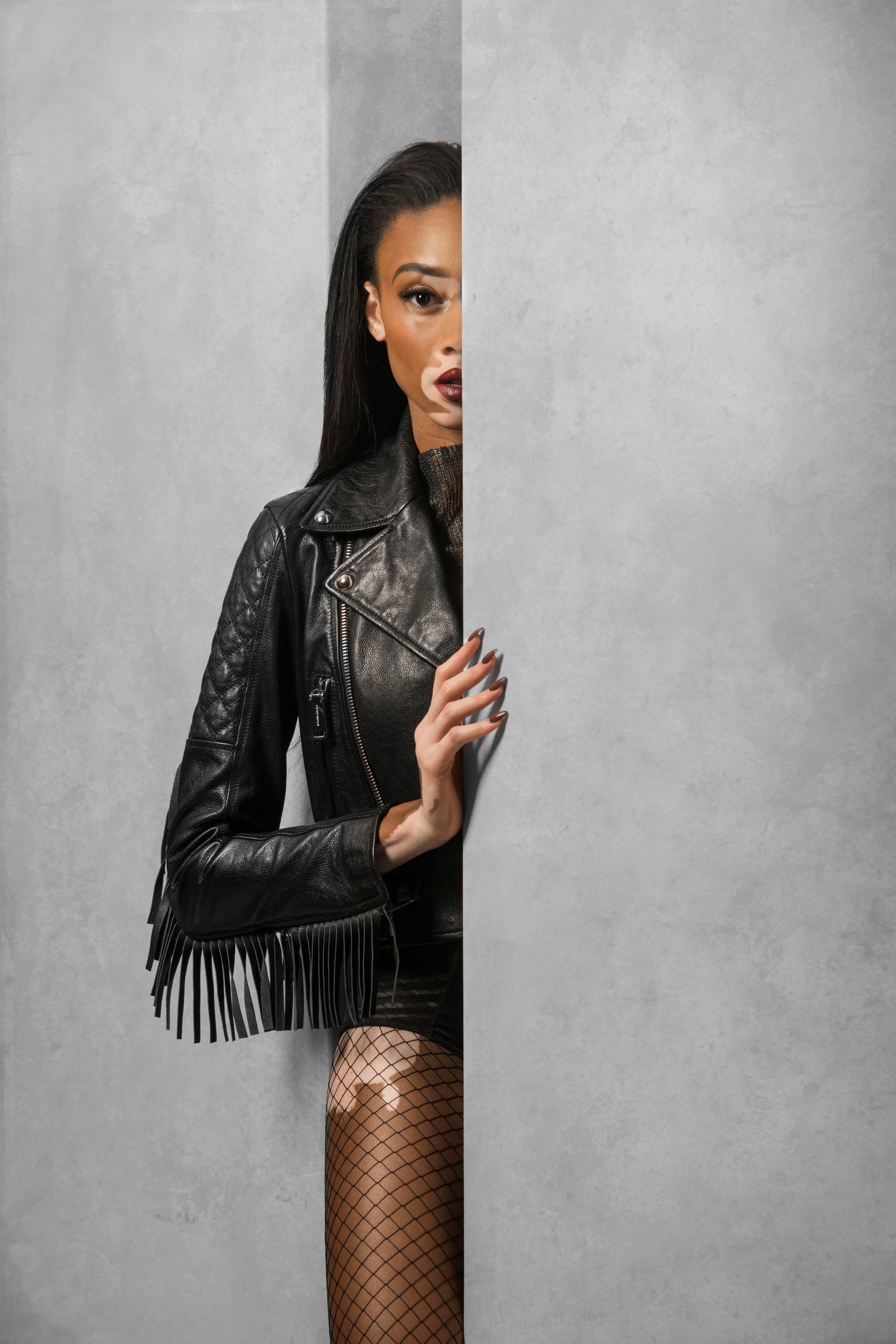A model in a leather jacket and fishnet tights peers around a concrete wall