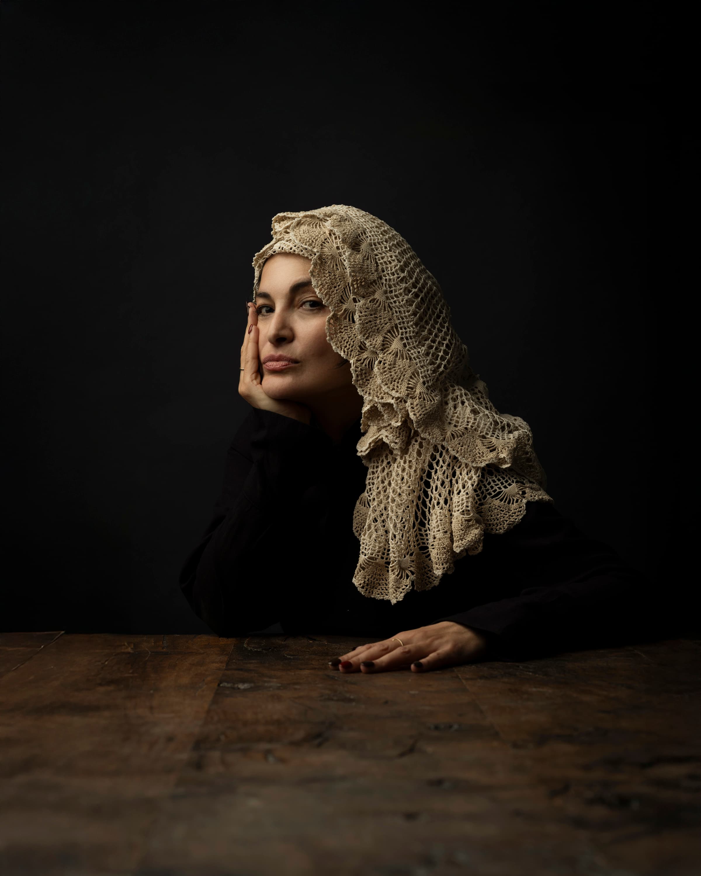 A woman in a lace headpiece rests her cheek on her hand at a dark wooden table
